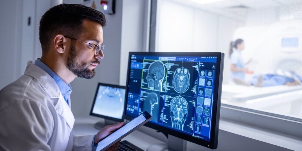 doctor examining X-ray images on display in MRI control room while in background nurse preparing the patient for examination test
