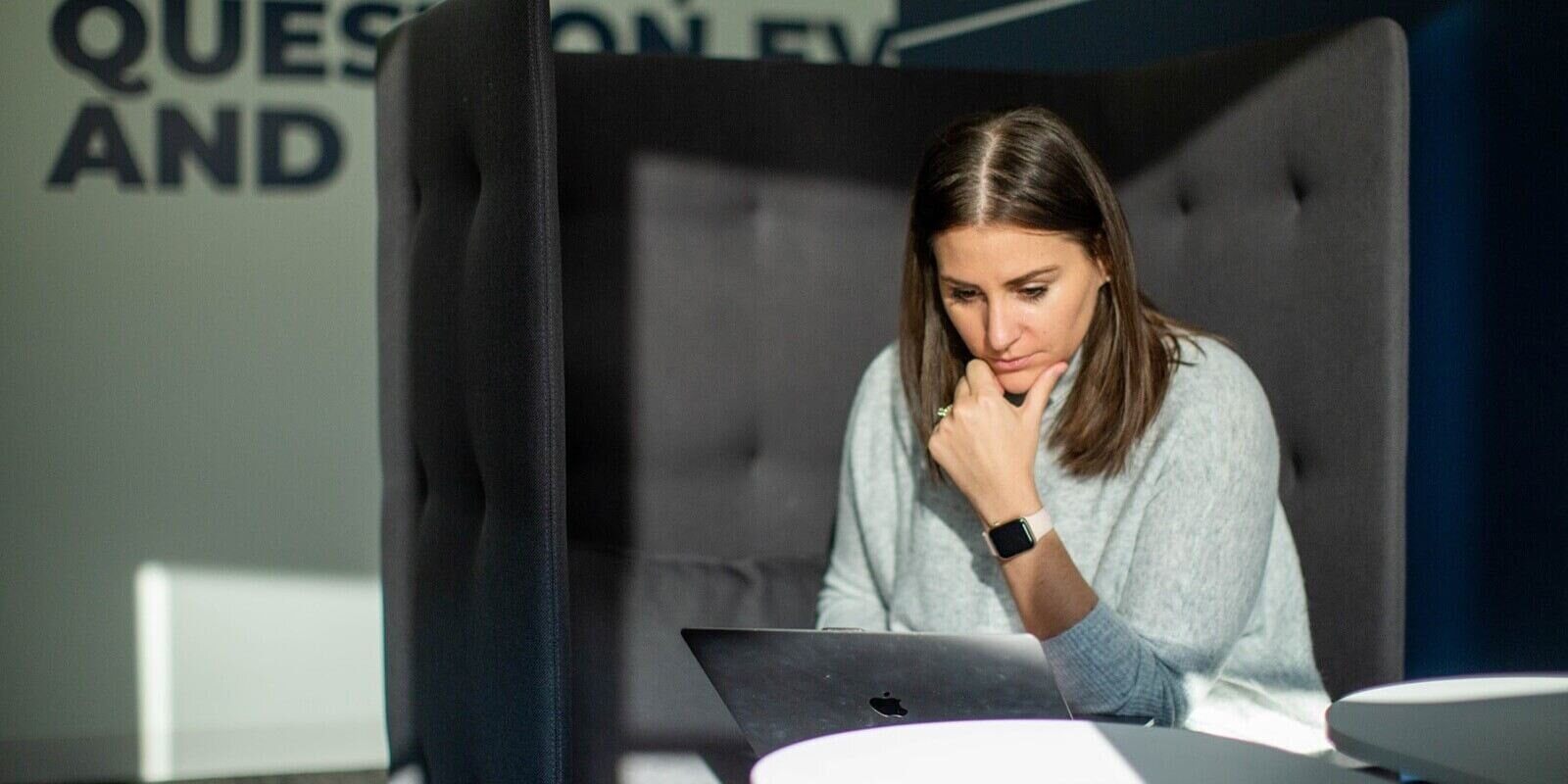woman sitting in a modern office booth, focused on her laptop, with sunlight casting shadow